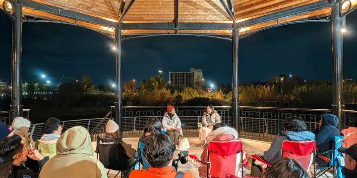 Di sera, il pubblico si riunisce al Music Pavilion aperto di Calgary; al centro, due persone sedute condividono racconti.