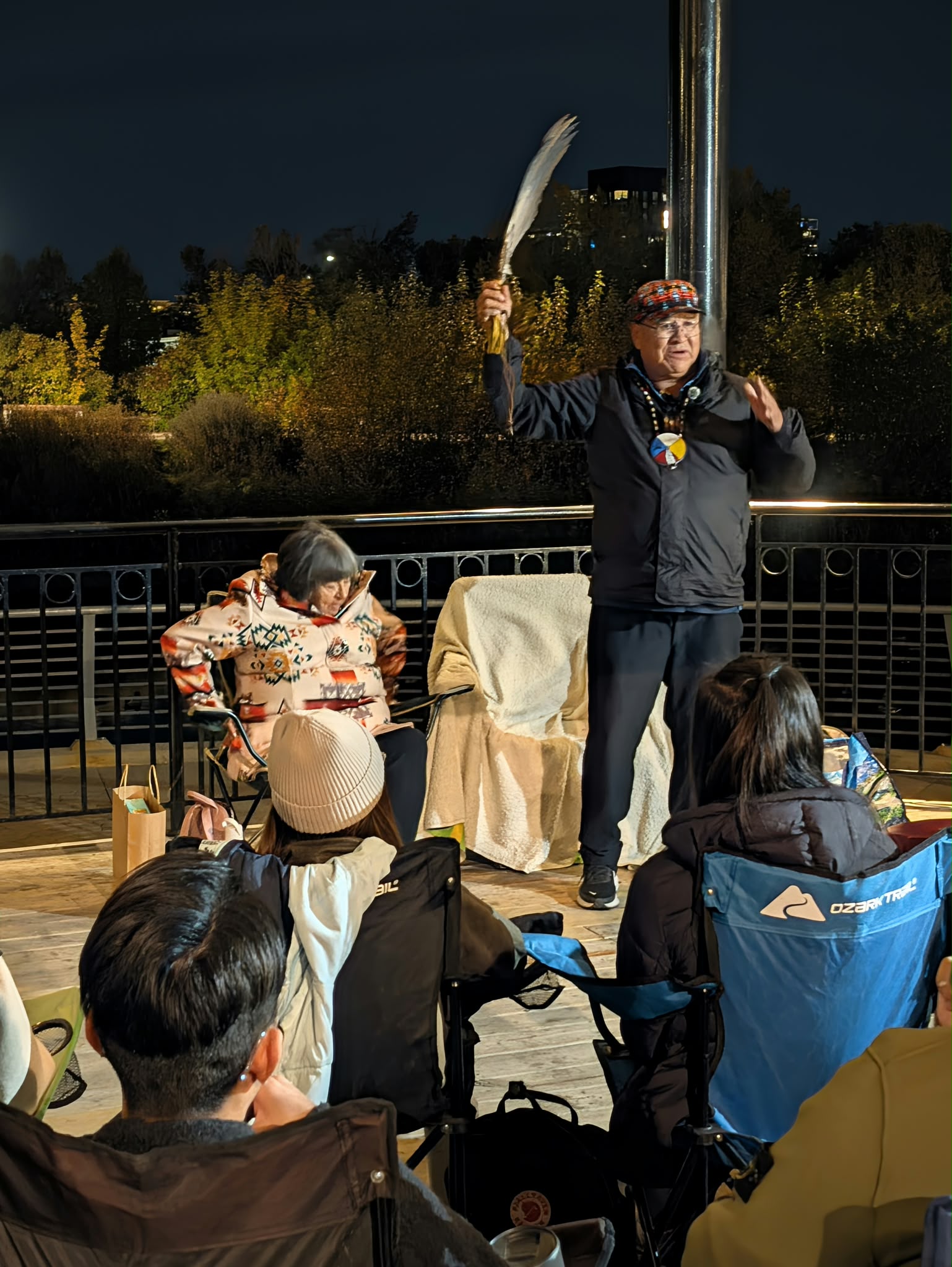 밤의 야외 파빌리온에서 한 연사가 깃털을 든 채 청중에게 이야기하고 있다.
